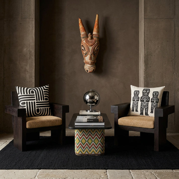 Photo professionnelle de décoration d'intérieur, salon afro chic brutaliste, vue de face. Deux fauteuils sculpturaux bas en bois de wengé massif aux lignes géométriques monolithiques, assises tissu texturé sable, chacun est orné d'un large coussin mzelle design motifs tribal noir . Murs en béton brut teinté argile sombre décoré d'un masque en bois peint .Table basse au design artisanal socle en bois et perles.