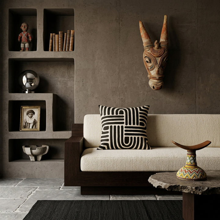 Photo professionnelle de décoration d'intérieur, salon afro chic brutaliste, vue de face. Canapé sculptural bas en bois de wengé massif aux lignes géométriques monolithiques, assises tissu texturé sable, orné d'un large coussin mzelle design, motifs tribaux. Murs en béton brut teinté argile sombre, niches architecturales asymétriques intégrées exposant divers objets décoratifs. 