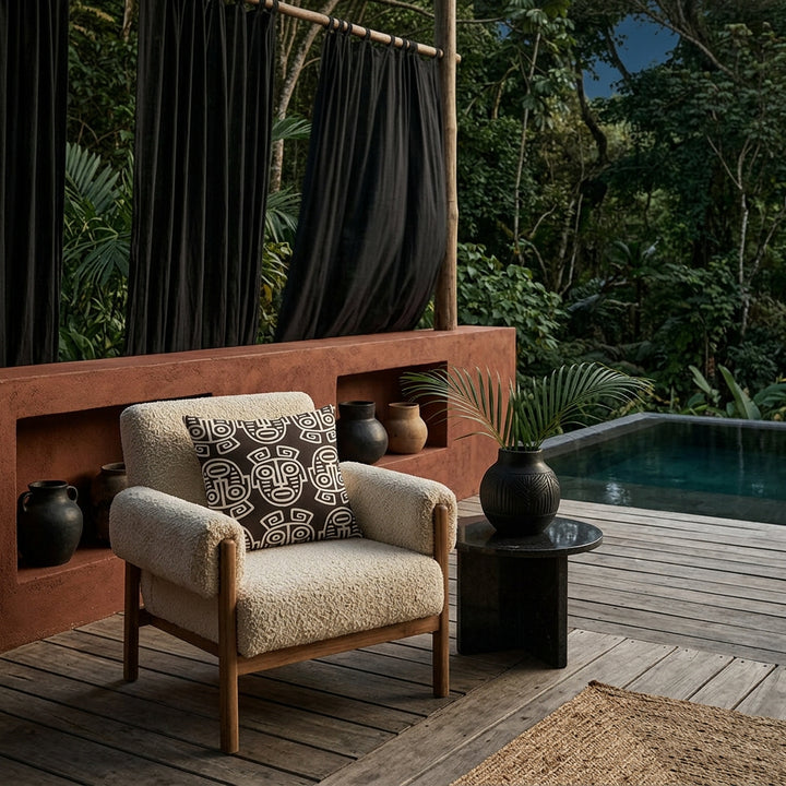 Photo professionnelle lifestyle, décoration terrasse d’eco-lodge du costa-rica, grand fauteuil en tissu crème texturé orné d'un large coussin mzelle design motif masques maya, table basse en pierre volcanique, sol en bois brut, poteries artisanales dans un muret peint en rouille, rideaux en lin noir flottant dans une brise chaude, architecture ouverte sur une jungle luxuriante et piscine sombre moderne.
