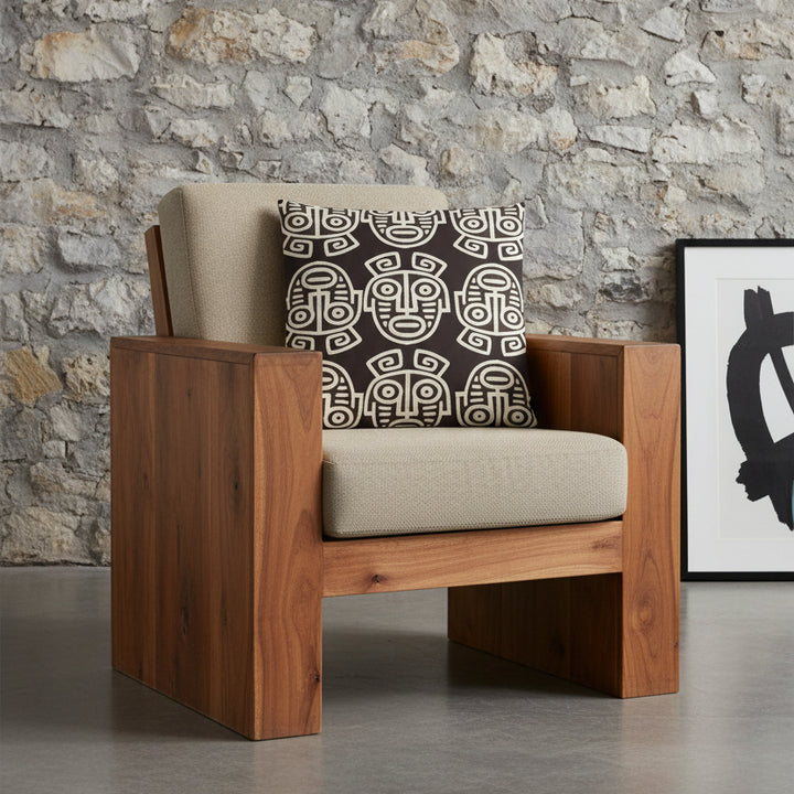 Photographie professionnelle studio de décoration haut de gamme : un coussin carré disposé sur un fauteuil brutaliste en bois de manguier, dossier et assise en twill beige, disposé contre un mur de pierres naturelles dans un décor neutre.
