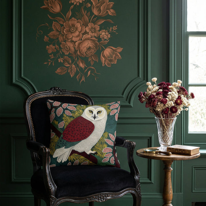 photographie professionnelle,  boudoir maximaliste italien, atmosphère intime et théâtrale , vue de face sur une chauffeuse régence en velours noir placée contre une fenêtre, où est posé un large coussin mzelle design motif chouette vintage. Mur vert profond avec moulures classiques, partiellement décoré d'une fresque florale baroque cuivrée, guéridon en bois de chêne patiné et bouquet de Fleurs .