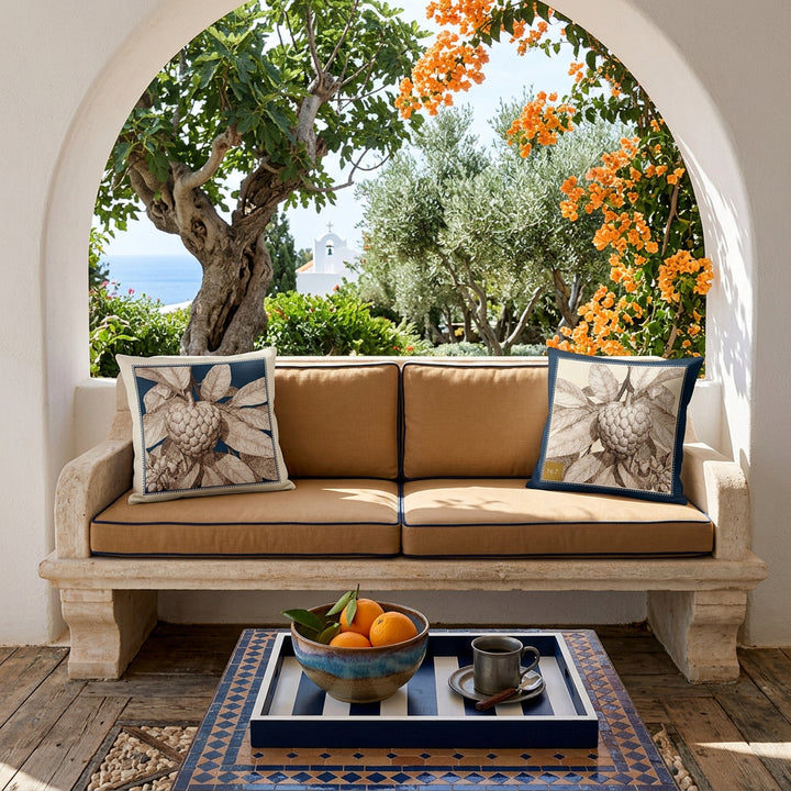 Photo professionnelle de décoration  d'une villa grecque baignée de soleil , banquette 2 places taillée dans la pierre blanchie , assises camel ornée de chaque côté d'un large coussin mzelle design motif attier, table basse recouverte de mosaiques bleu et brun, plateau en bois garni d'une coupe en grès marron dégradé contenant trois oranges, une tasse de café, vue sur jardin et mer.