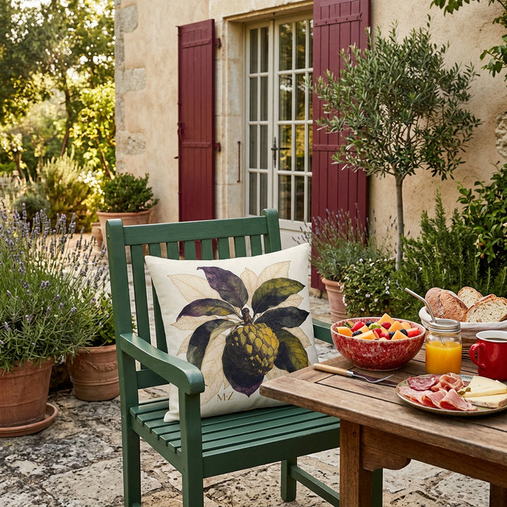 Photographie professionnelle terrasse provençale, une chaises en bois peint vert foncé, ornée d'un beau coussin mzelle design motif tropical attier. A côté d'une Table rustique dressée avec déjeuner estival gourmand. En arrière plan la maison et sa grande baie vitrée, buisson de lavande, un oliviers, jardin estival.
