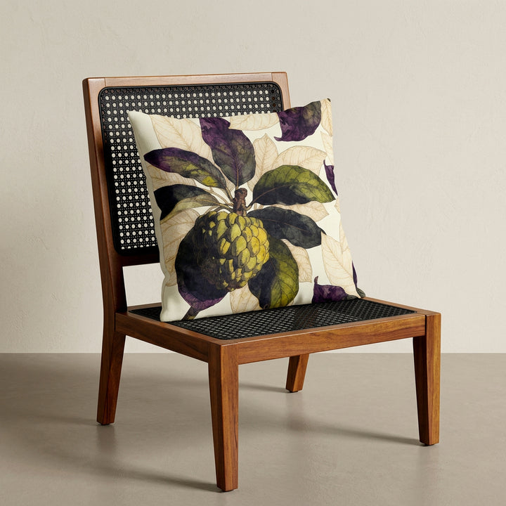 Photographie d'un grand coussin carré mzelle design motif fruit tropical attier, disposé sur une chaise créole en bois de manguier verni, sans accoudoirs, dossier et assise en bois arrondi demi lune, dans décor écru neutre.
