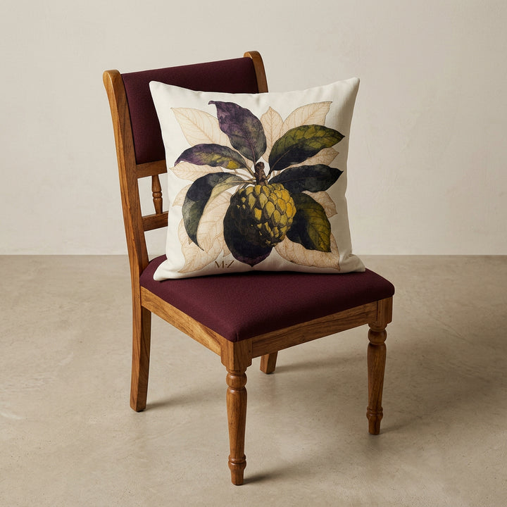 Photographie d'un grand coussin carré mzelle design motif fruit tropical attier, disposé sur une chaise créole en bois de manguier verni, sans accoudoirs, dossier et assise en bois arrondi demi lune, dans décor écru neutre.
