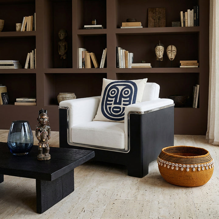 Photo professionnelle lifestyle, décoration Salon ethno-brutaliste , imposant fauteuil en lin blanc , liseré chrome , orné d'un large coussin mzelle design motif pétroglyphe. Table basse en bois brûlé noir garni d'un vase en verre fumé bleu et d'une statuette style vaudou artistique, architecture minérale et mur peint en brun cacao avec niches de bibliothèque.