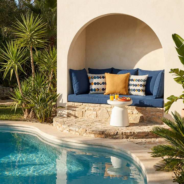 Photo professionnelle de décoration villa provençale, vue de la piscine arrondie qui se termine à la frontière d'une alcôve en arche, où est niché un canapé taillé dans la pierre surélevé sur une marche arrondie en pierres rustiques, assises en twill bleu marine avec un large coussin ocre au centre, autour 2 coussins larges mzelle design , et une table d'appoint en résine blanche garnie de 2 verres de jus de fruit et tapas, jardin tropicalisé autour, ambiance estivale invitante.