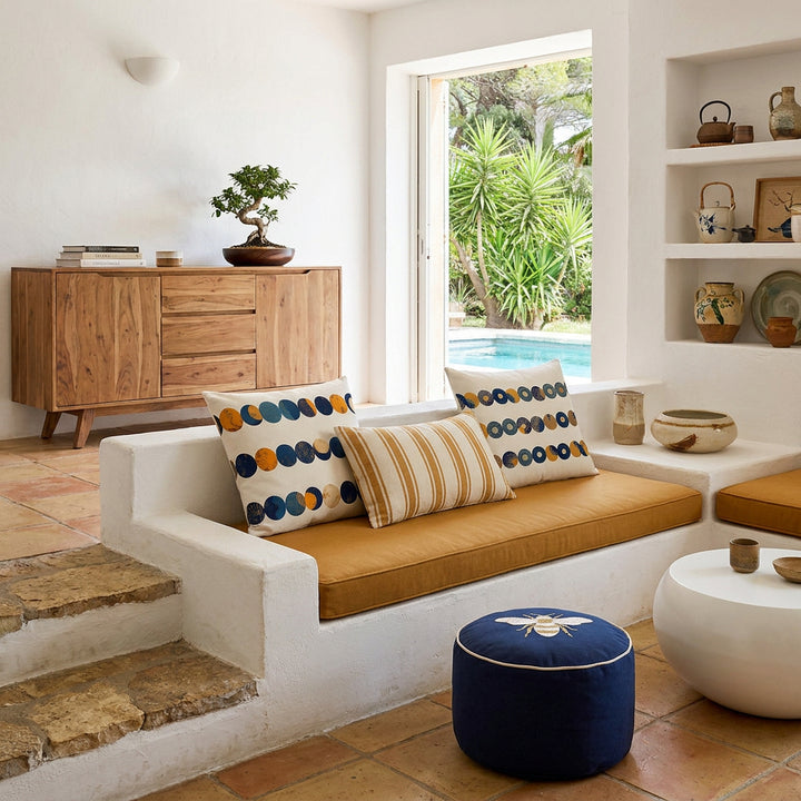 Photo professionnelle de décoration d'intérieur, salon Arty dans une villa provençale, vue de face d'un canapé taillé dans la pierre blanchie surélevé sur une marche en pierres rustiques, assises en twill ocre avec un coussin rectangle rayé écru/ocre au centre, autour 2 coussins larges mzelle design motifs minimalistes, et un petit pouf en coton bleu marine brodé d'une abeille blanc et ocre.