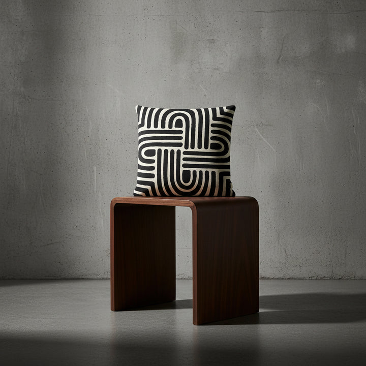 Photographie professionnelle studio,  de haute qualité : un coussin carré mzelle design motif graphique noir et blanc, disposé sur un tabouret en bois noir de wengé, design moderne, disposé contre un mur brutaliste, dans un décor neutre.