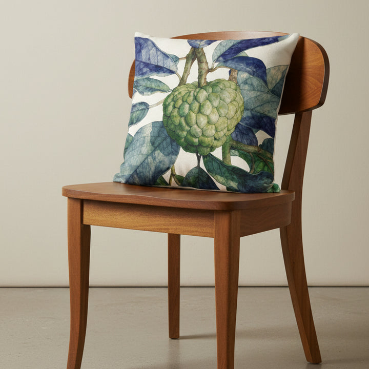 Photographie d'un grand coussin carré mzelle design motif fruit tropical attier, disposé sur une chaise créole en bois de manguier verni, sans accoudoirs, dossier et assise en bois arrondi demi lune, dans décor écru neutre.