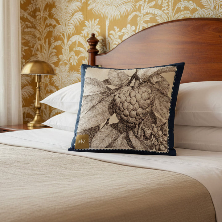 Photographie décoration chambre esprit créole vue de face d'un grand coussin carré mzelle design motif fruit tropical attier, tête de lit arrondie demi lune, murs avec papier peint ocre et blanc motif jungle, voilage, lampe en laiton, draps en coton blanc et lin naturel beige.