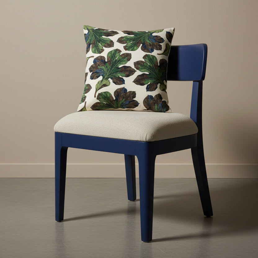 Photographie professionnelle vue de 3/4, extrêmement détaillée et de haute qualité d'un grand coussin  mzelle design motif feuilles all over, disposé sur une chaise moderne de forme arrondie, en bois peint bleu indigo, sans accoudoirs, dossier et assise en tweed écru, disposé dans décor beige neutre.