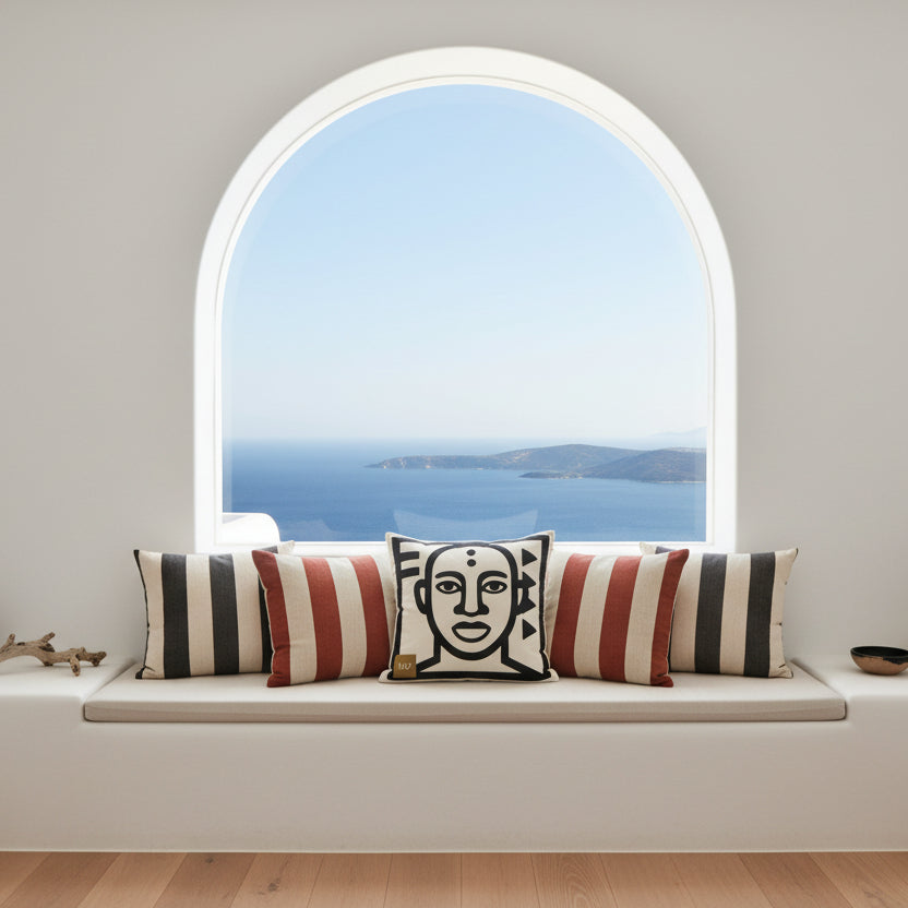 Photographie professionnelle d'une villa cycladique, une banquette moderne en maçonnerie, orné au milieu le large coussin carré, et 3 coussins à rayures dans les tons noir, rouille et terre autour.  atmosphère accueillante.