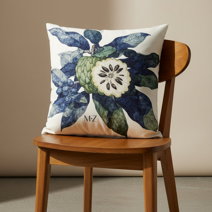 Photographie d'un grand coussin carré mzelle design motif fruit tropical attier, disposé sur une chaise créole en bois de manguier verni, sans accoudoirs, dossier et assise en bois arrondi demi lune, dans décor écru neutre.