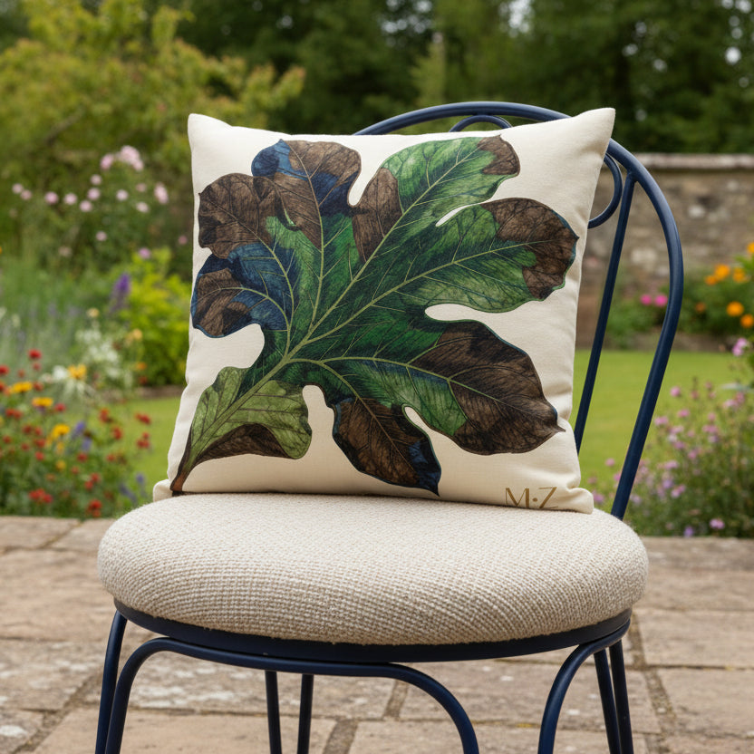Photographie professionnelle vue de 3/4, extrêmement détaillée et de haute qualité d'un grand coussin mzelle design motif feuille, disposé sur une chaise en fer forgé noire de forme arrondie, disposée sur une terrasse à la campagne, vue sur jardin.