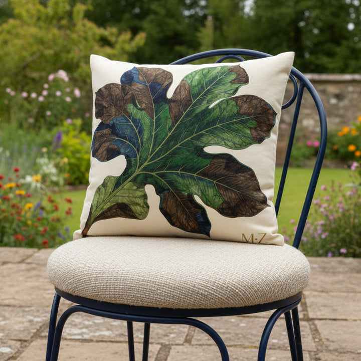 Photographie professionnelle vue de 3/4, extrêmement détaillée et de haute qualité d'un grand coussin mzelle design motif feuille, disposé sur une chaise en fer forgé noire de forme arrondie, disposée sur une terrasse à la campagne, vue sur jardin.