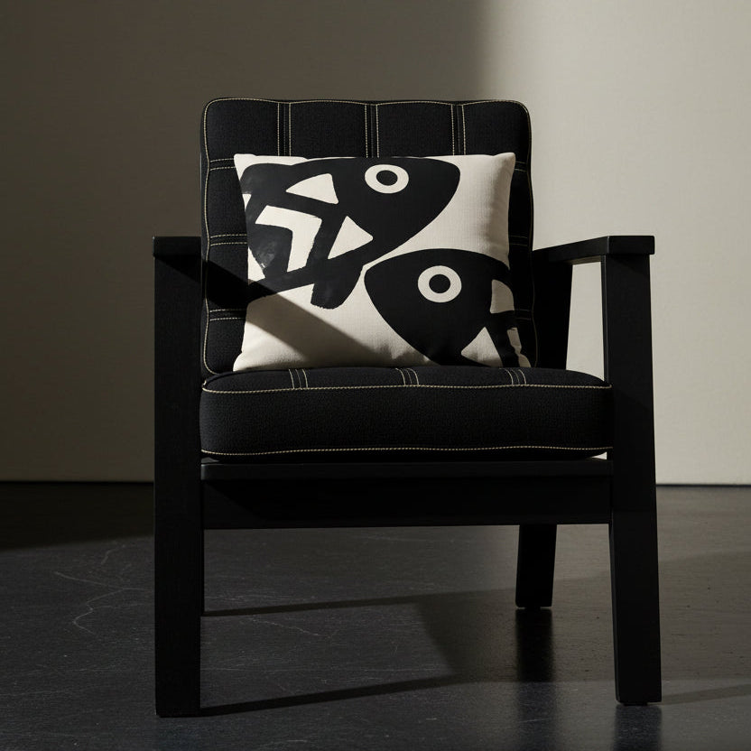 Photographie professionnelle qualité studio, d'un coussin carré mzelle dsign motifs poissons disposé sur une chaise japonaise sans accoudoirs, dossier en tissu noir surpiqué de fil écru, structure épaisse, disposé dans décor écru neutre, sol en béton ciré noir.